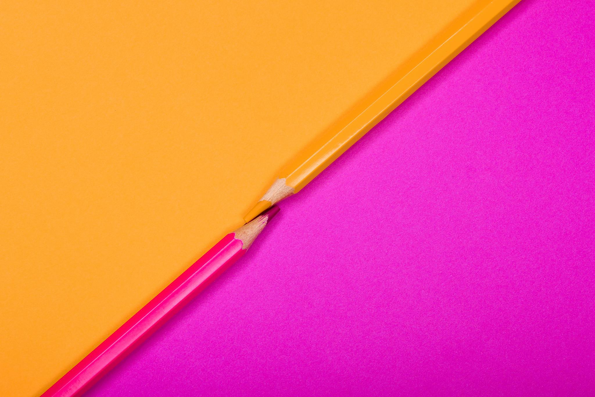 Two colored pencils on bright pink and yellow backgrounds, creatively arranged.